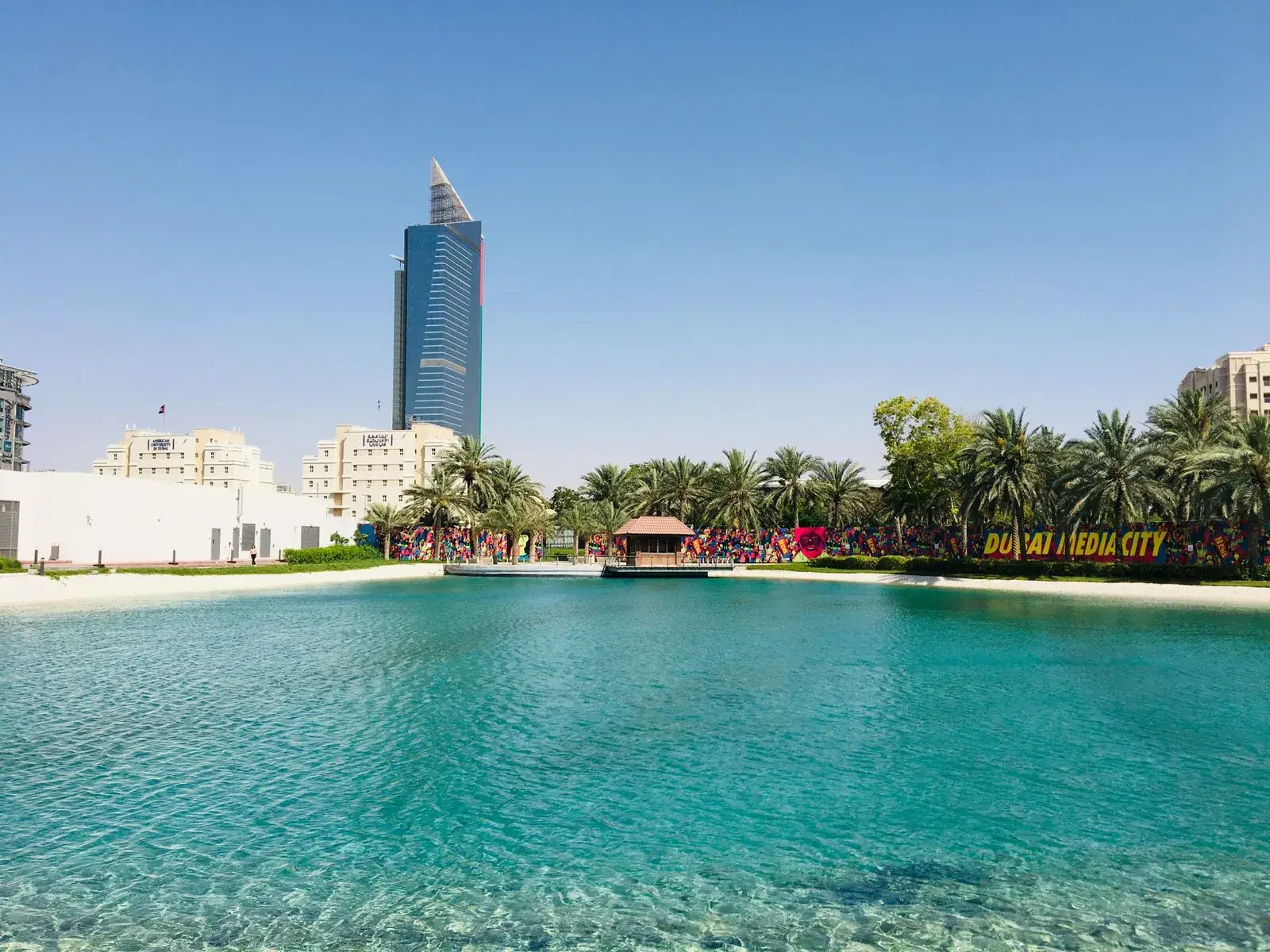 DUBAI MEDIA CITY painted across a wall behind a turquoise lagoon and palms, a tall blue tower rising behind it.